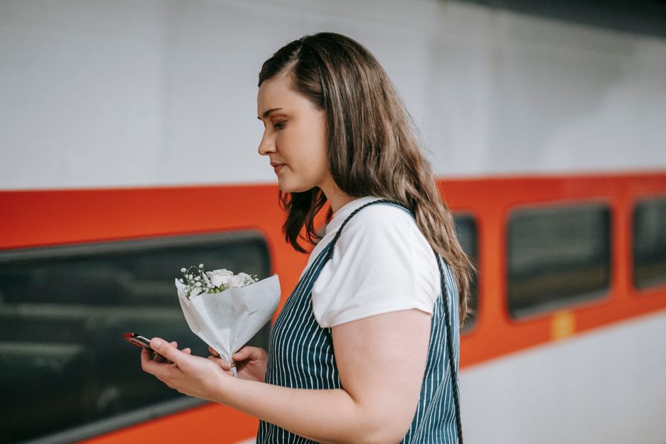 A young woman with long, wavy brunette hair holding a small floral bouquet wrapped in white paper, featuring white roses and green foliage, standing on a platform near a red train. She is dressed in a
